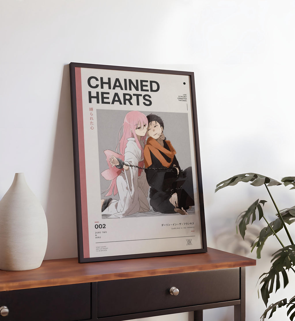 a framed poster of two anime characters, one with pink hair and the other with dark hair, sitting together and appearing to embrace. The poster is placed on a wooden table, and there is a plant in the background.