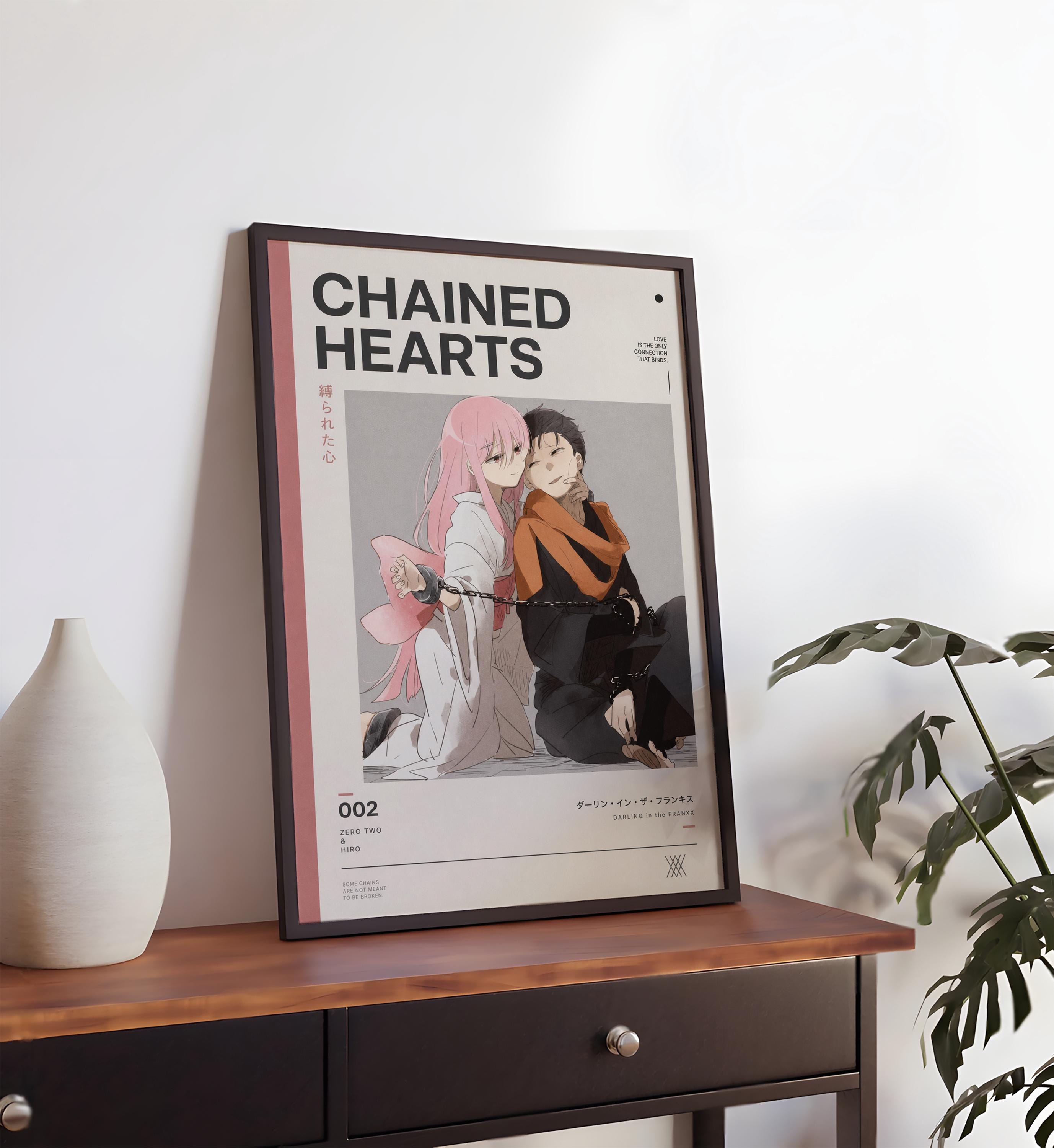 a framed poster of two anime characters, one with pink hair and the other with dark hair, sitting together and appearing to embrace. The poster is placed on a wooden table, and there is a plant in the background.